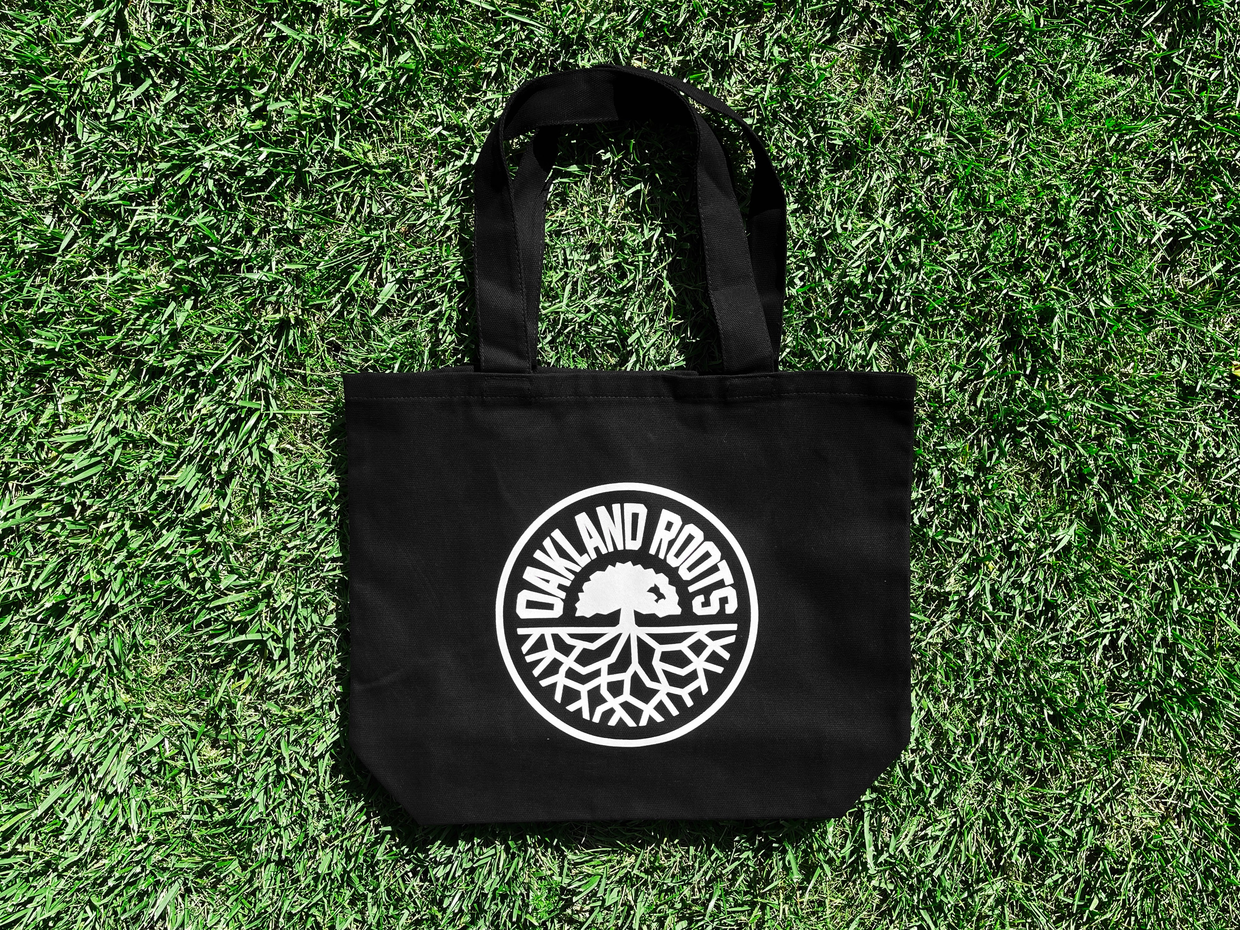 Black reusable shopping tote with large white Oakland Roots logo crest lying on grass.