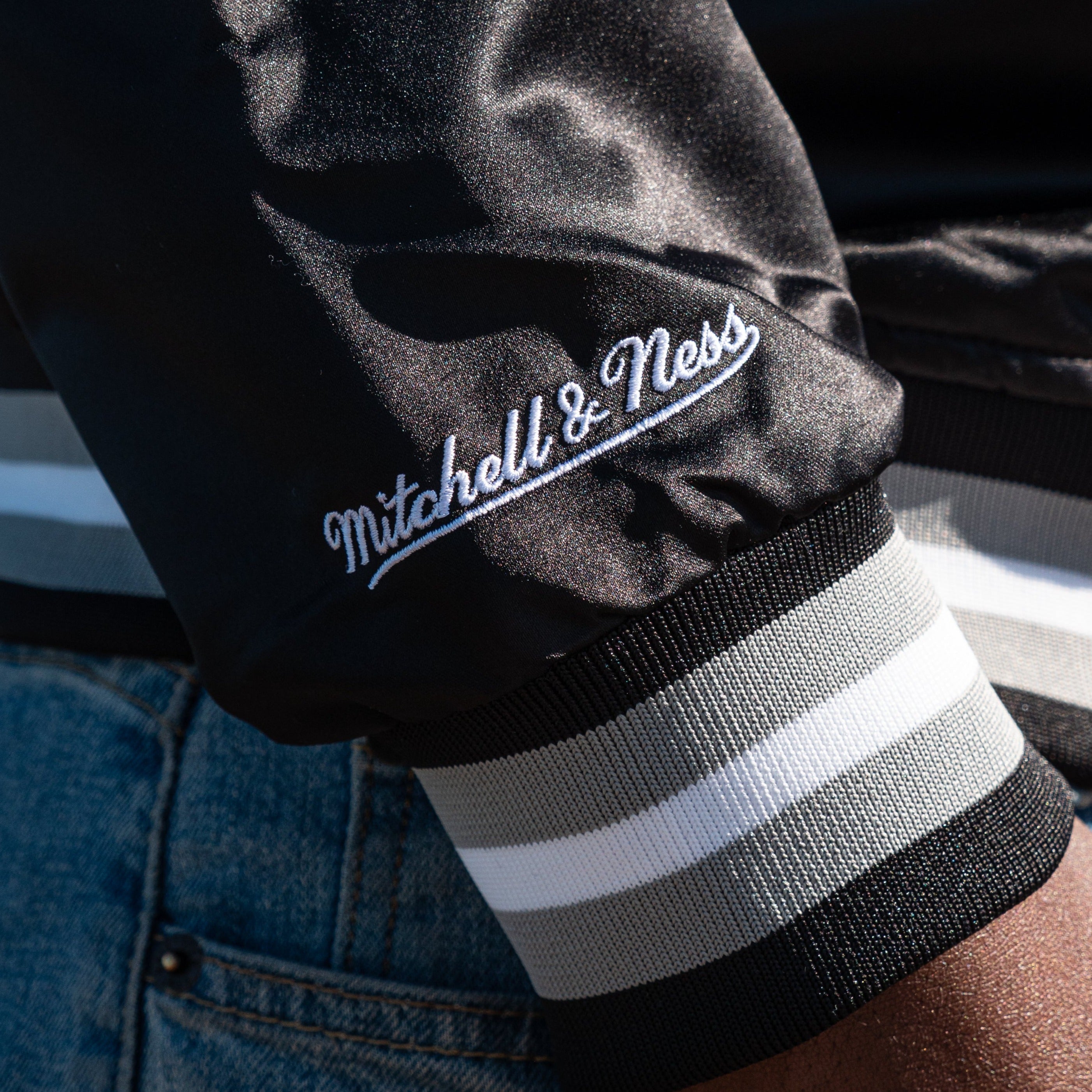 Close-up of a person’s arm wearing the Mitchell & Ness Oakland Roots SC Lightweight Satin Jacket with "Mitchell & Ness" embroidered in white, cursive text. The jacket from Oakland Roots SC features a ribbed cuff with gray, black, and white stripes. The person is also wearing blue denim jeans.