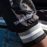 Close-up of a black satin jacket sleeve with embroidered Mitchell Ness wordmark.