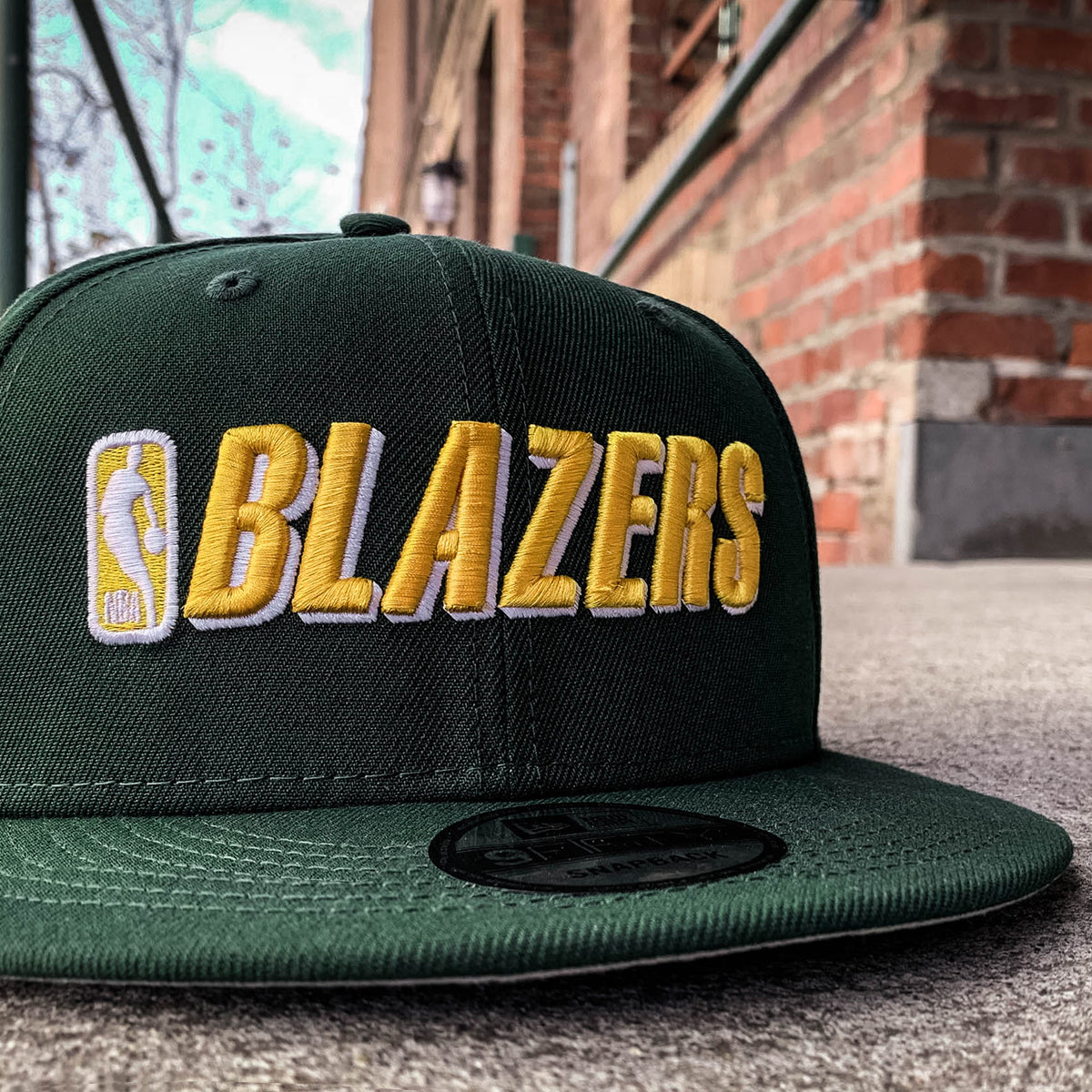 Green cap with embroidered gold NBA BLAZERS logo on the crown sitting outdoors on the street.