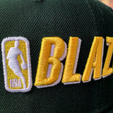 Close-up of gold NBA BLAZERS logo on the crown of a green cap.