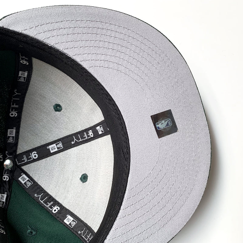 Grey inside the crown of a green cap with black taping with 9FIFTY New Era wordmarks on repeat.