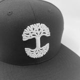 Close-up of silver embroidered Oaklandish tree logo on a black cap with black bill.