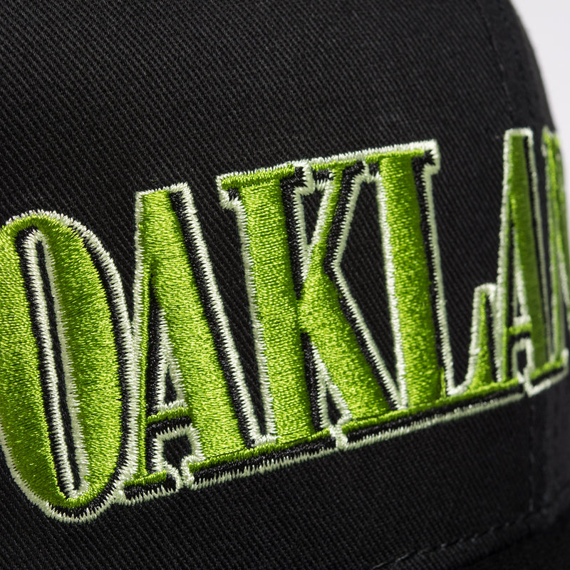 Close up of green embroidered Oaklandish tree logo on the front of a black cap.