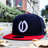 A navy New Era cap with a white embroidered A's O logo and a New Era sticker on the red bill on leafy Oakland street.