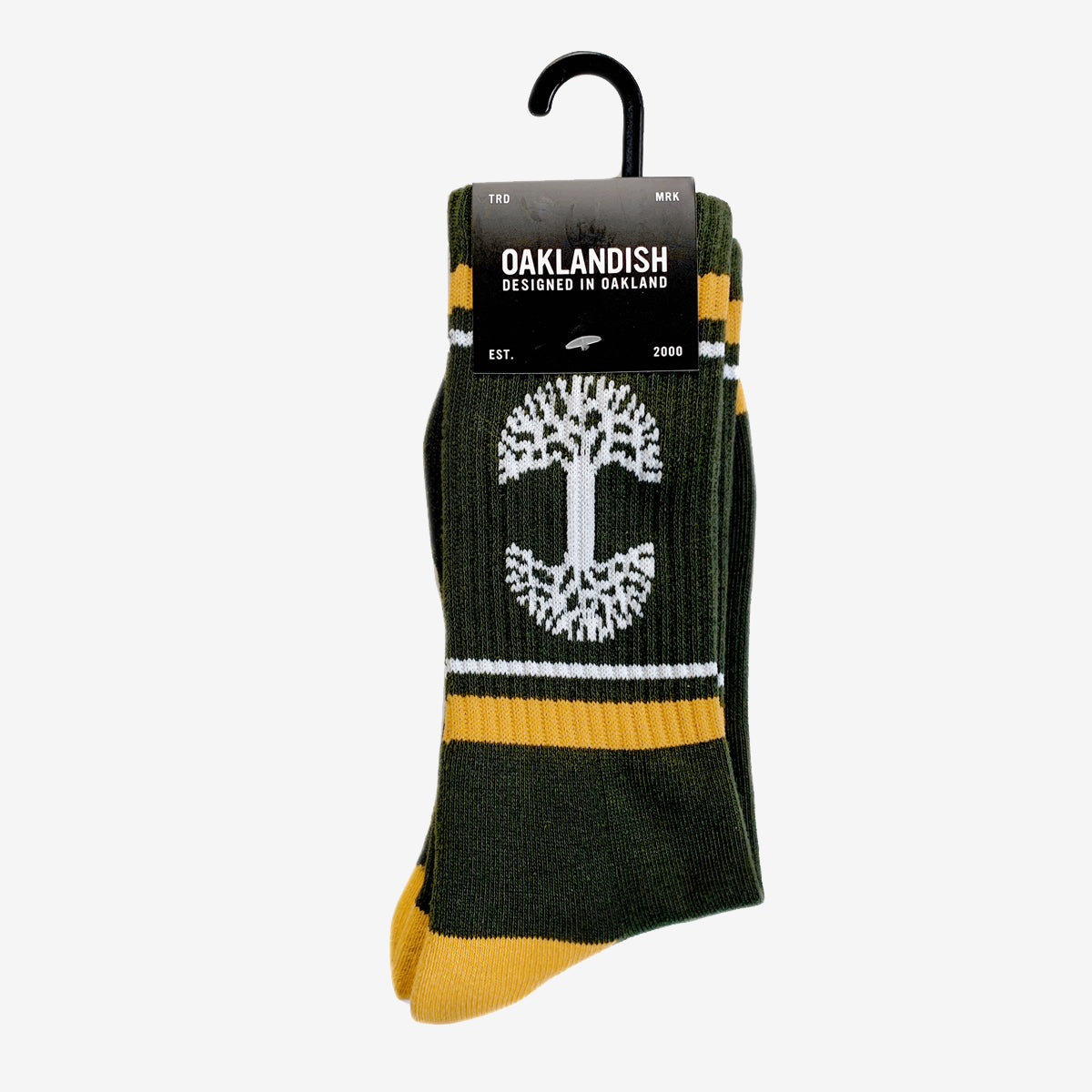 Introducing the OAK Knit Crew Socks by Oaklandish, a stylish pair crafted in forest green with yellow accents and adorned with a white tree design at the center. Made from a comfortable blend of cotton, polyester, and spandex, these crew socks also showcase the Oaklandish logo. They come packaged with a sleek black label that reads "OAKLANDISH - Designed in Oakland - Est. 2000," and include a convenient plastic hanger for easy display.