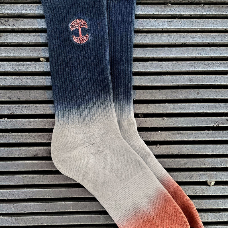 Two nestled high-cut, dip-dyed (navy, grey and pink) crew socks with an embroidered Oaklandish logo on a wood deck.
