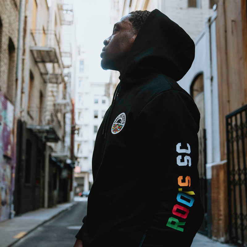 Man standing sideways on street wearing black Oakland Roots SC hoodie with color Roots SC wordmark on the sleeve.