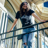 Women leaning against a railing, an ornate building in the background, wearing a black t-shirt with an Oakland neighborhood map.