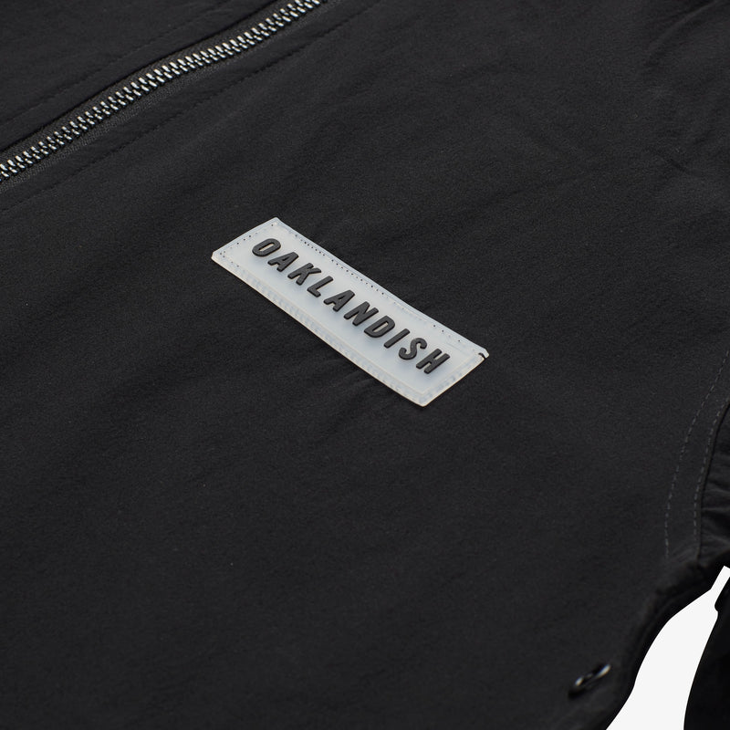 Close up of rubber patch with black OAKLANDISH wordmark on lightweight black zip up hooded jacket. 