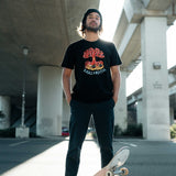 A man standing under an overpass with a skateboard with an Oaklandish DJ crew graphic on a black t-shirt.