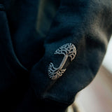 Close up of grey embroidered Oaklandish tree logo on the sleeve of a black hooded sweatshirt.
