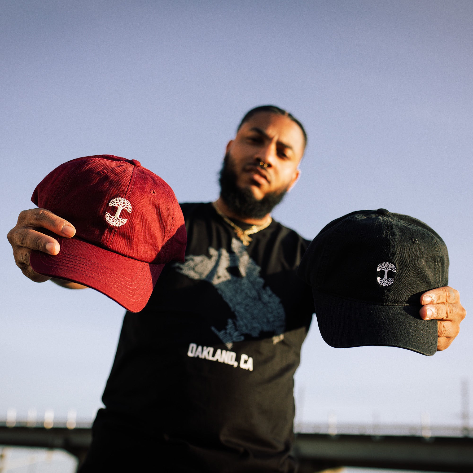 A man standing outside with arms stretched straight out with a red Oaklandish dad cap in one hand and a black one in the other.