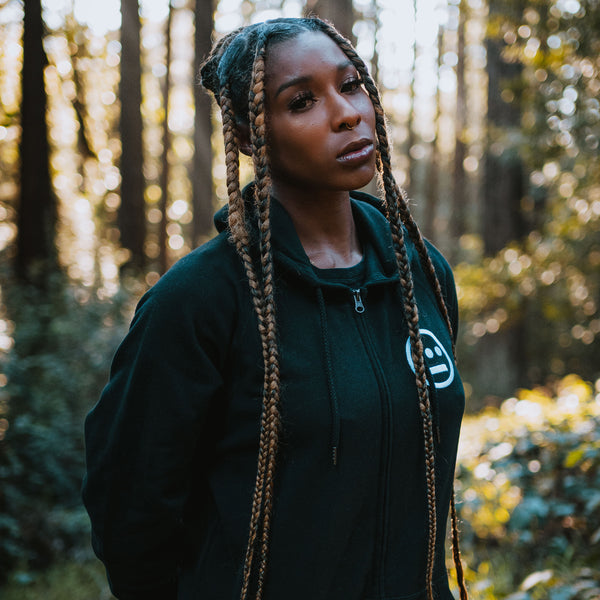 Woman standing in the forest wearing a black zip-up hoodie with white Hieroglyphics hip hop logo on the left chest wear side.