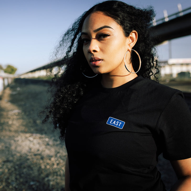 Women standing under train tracks in black t-shirt with word East in blue highway sign graphic on left breast.