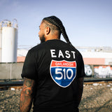 Man standing in train yard, back to camera, in black t-shirt with 510 East Oakland Interstate highway sign on back of shirt. 