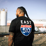 Man standing in train yard, back to camera, in black t-shirt with 510 East Oakland Interstate highway sign on back of shirt. 