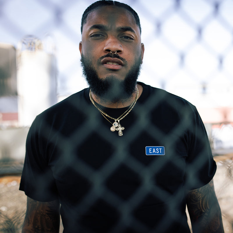 Man standing in front of chain link fence iin black t-shirt with word East in blue graphic on left breast to celebreate East Oakland.