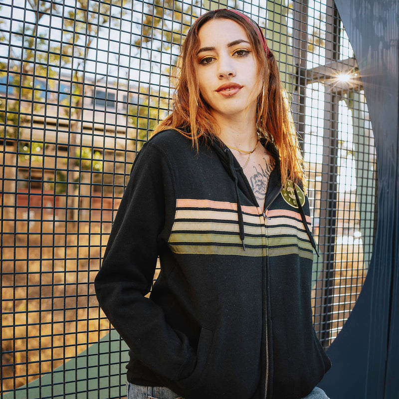 A female model wearing black Oaklandish zip-up hoodie with stripes, in front of metal structure.