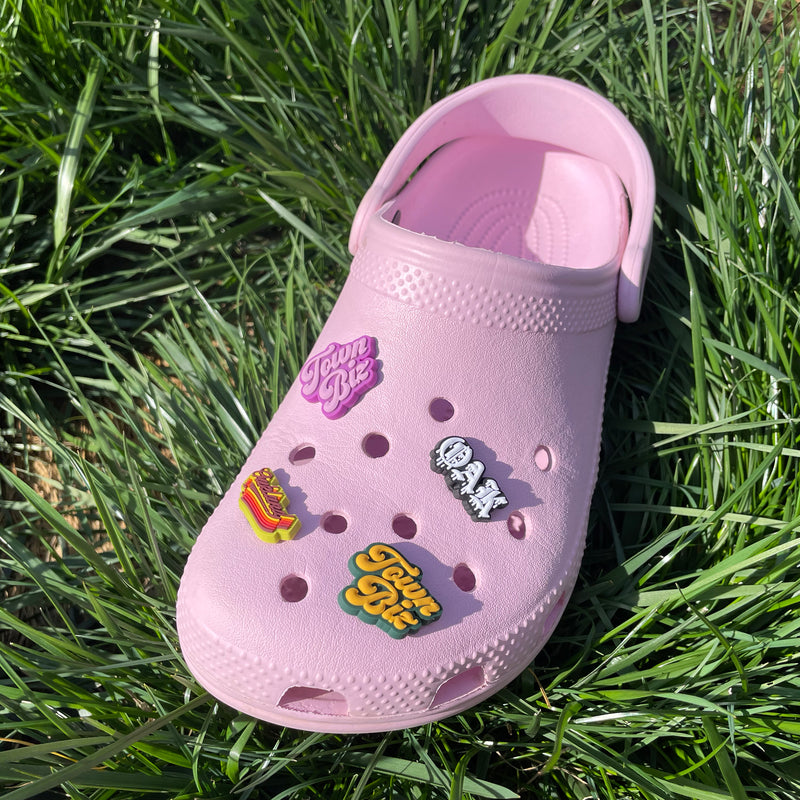 A single pink croc clog on grass decorated with three shoe charms - two Town Biz, Oak Bleed, Oakland Swash.