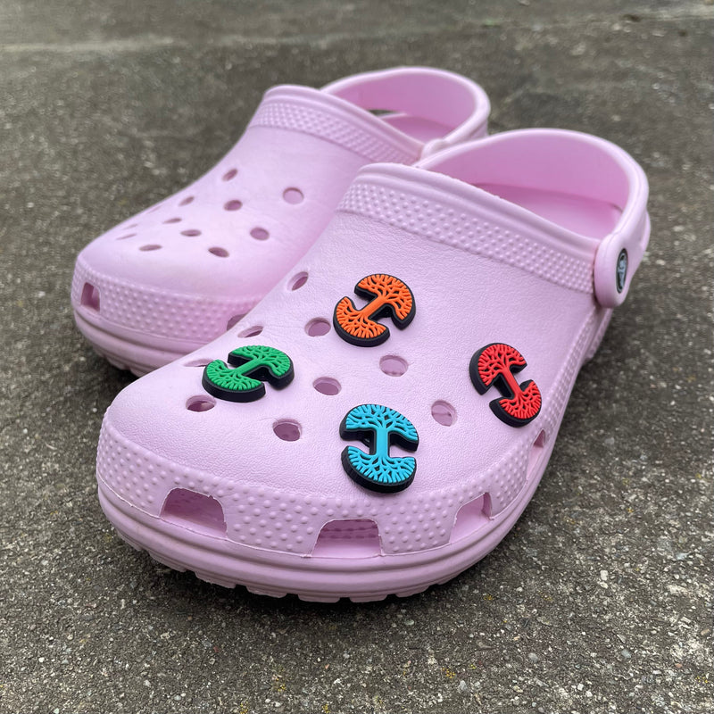 A pair of pink croc clogs on a sidewalk. The front one has four different color Oaklandish tree logo shoe charms.