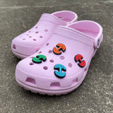 A pair of pink croc clogs on a sidewalk. The front one has four different color Oaklandish tree logo shoe charms.