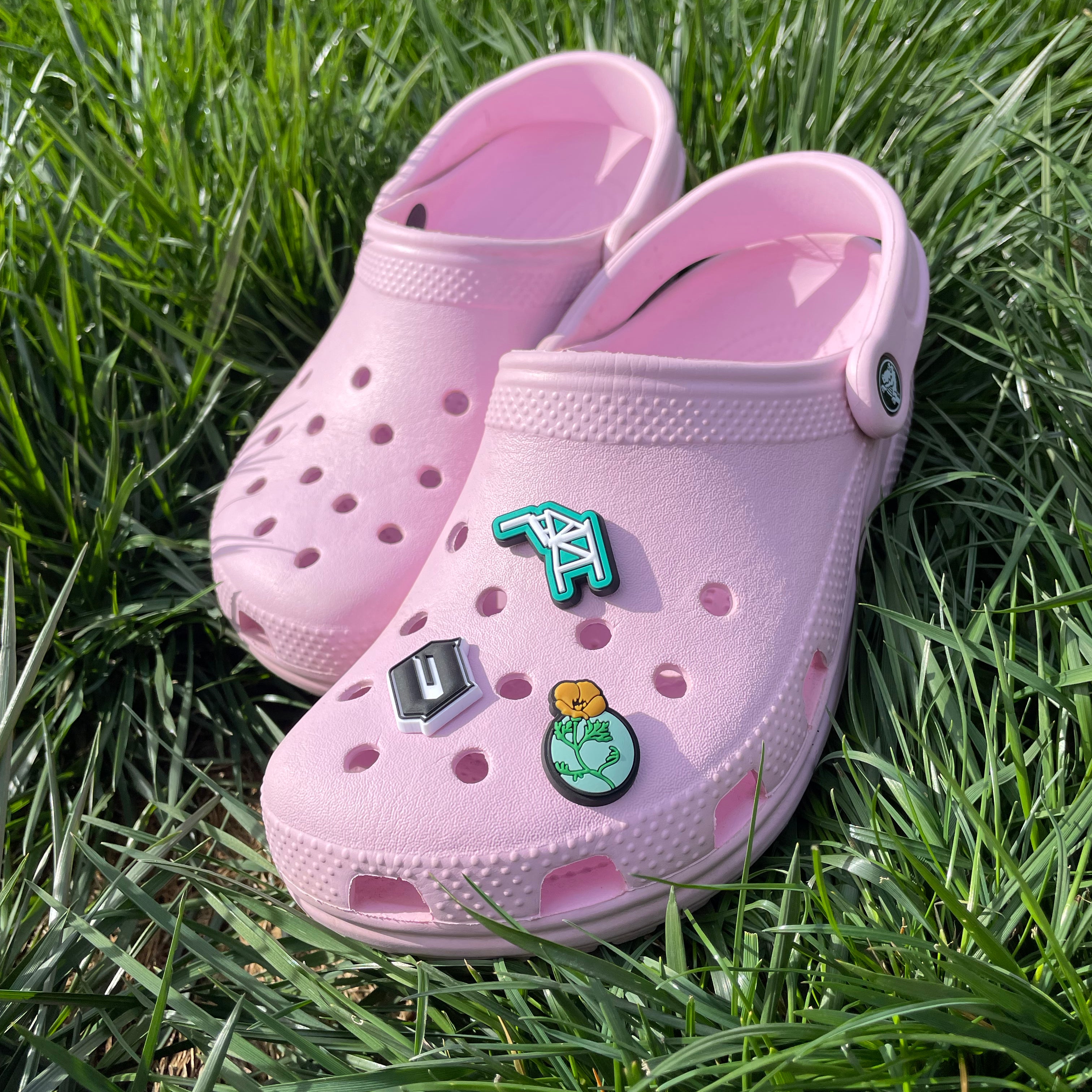 A pair of pink croc clogs on grass. The front one has three shoe charms - Cranes, California Poppy and Oaklandish O.