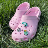 Two pink crocs sitting on the grass. One in the foreground is decorated with three shoe charms.
