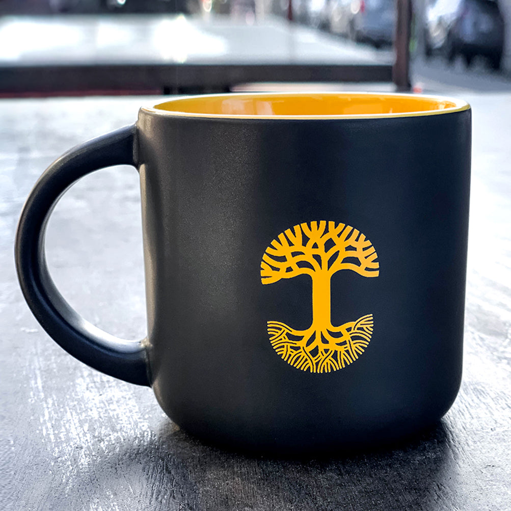Black ceramic mug with yellow inside with yellow Oaklandish tree logo on outdoor bench.