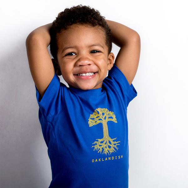 Smiling toddler wearing a royal blue toddler-sized t-shirt with yellow Oaklandish tree logo and wordmark on the chest.