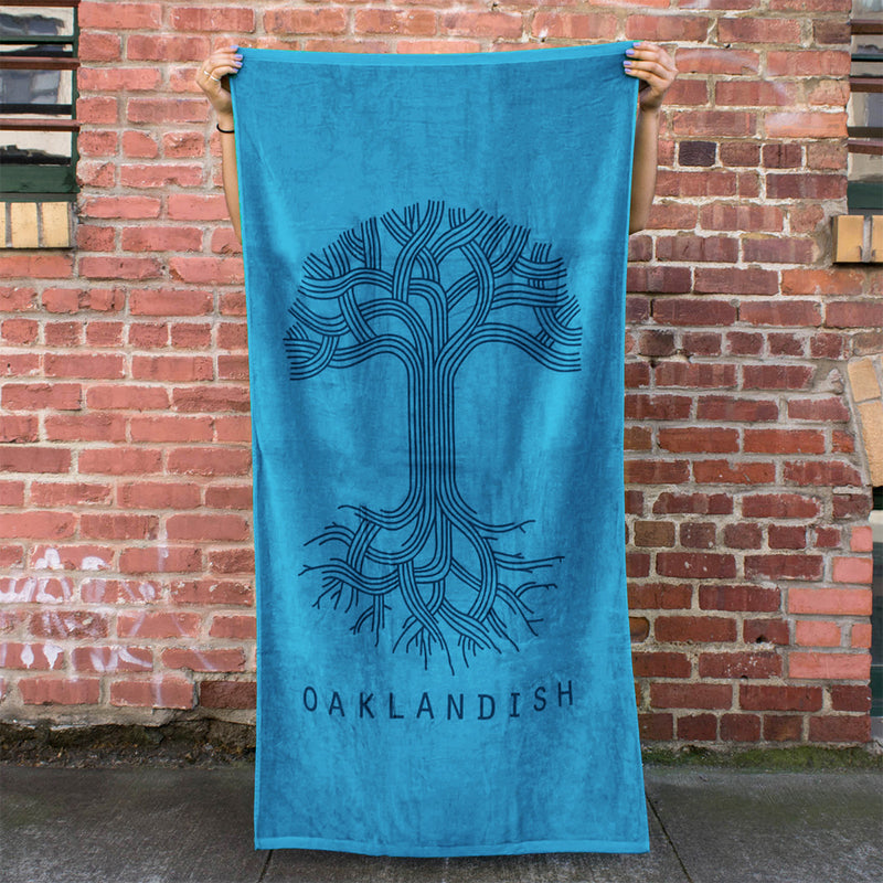 A person standing outside holding a plush oversized beach towel in aqua blue with a classic Oaklandish logo and wordmark.