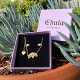 Necklace with three Oakland trees and heart with Chula Aesthetics wordmark in purple Chula jewelry box in an outdoor planter.