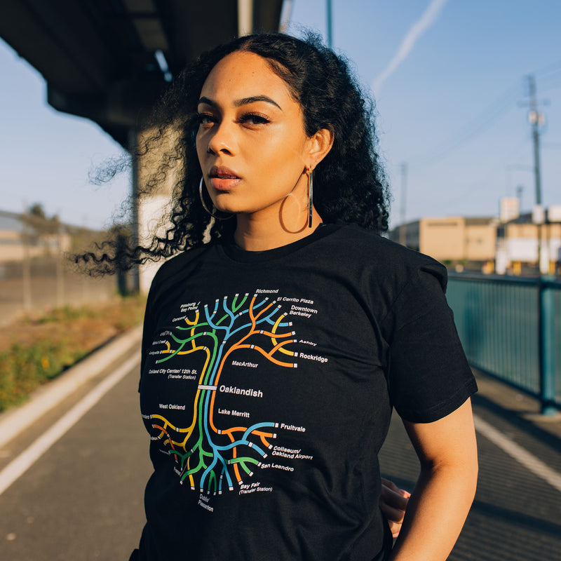Woman standing under an overpass wearing a black t-shirt with a BART transit map in the shape of the Oaklandish tree logo.