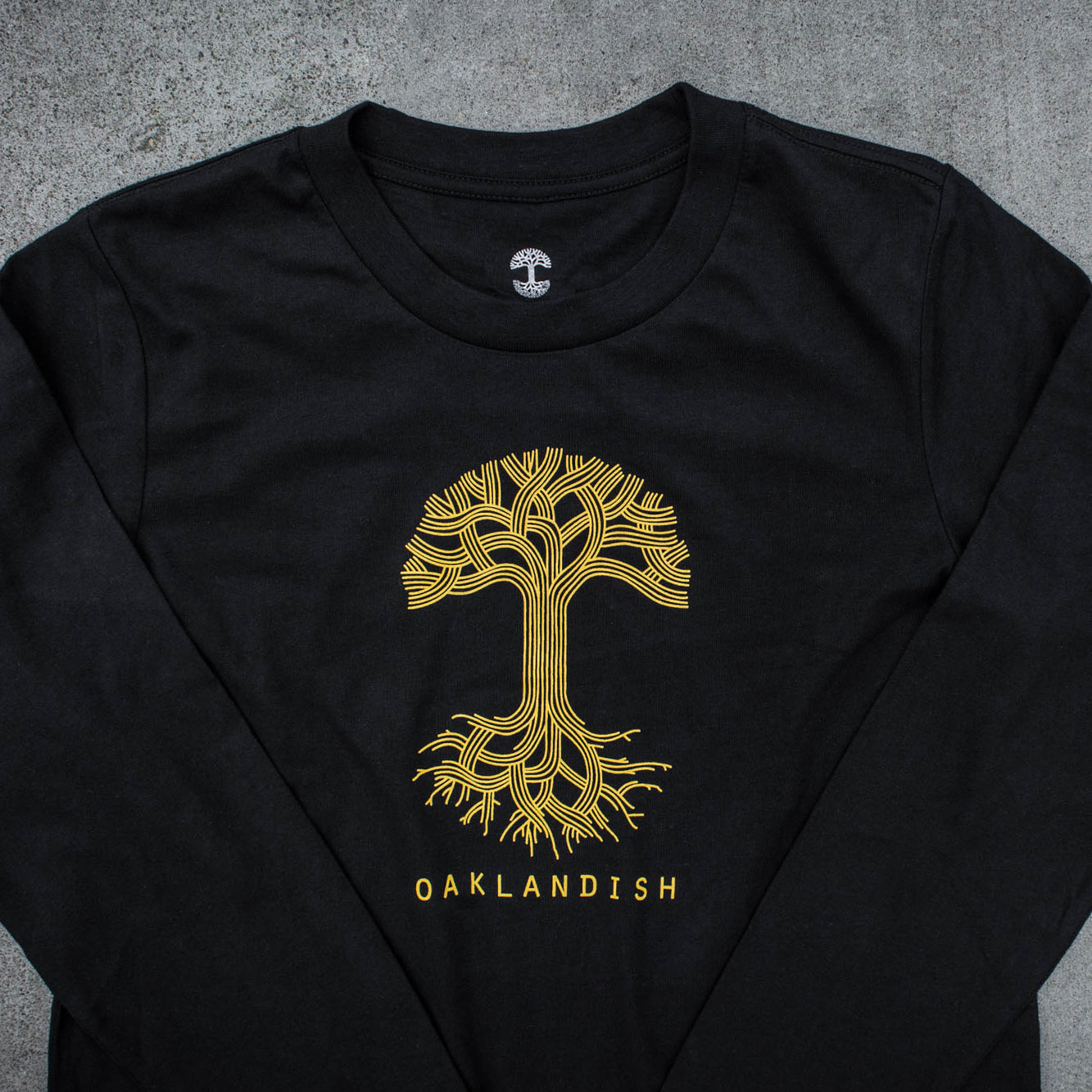 Close-up of gold Oaklandish tree logo and wordmark on the chest of a women’s cut black long-sleeve t-shirt with arms folded.