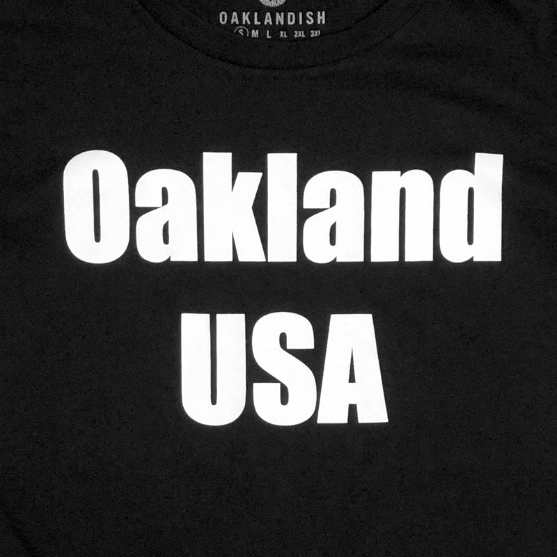Close-up of a large white Oakland USA wordmark logo on the chest of a black women’s t-shirt.