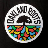Close-up of a full-color Oakland Roots circle logo on the chest of a women’s black tank-top.