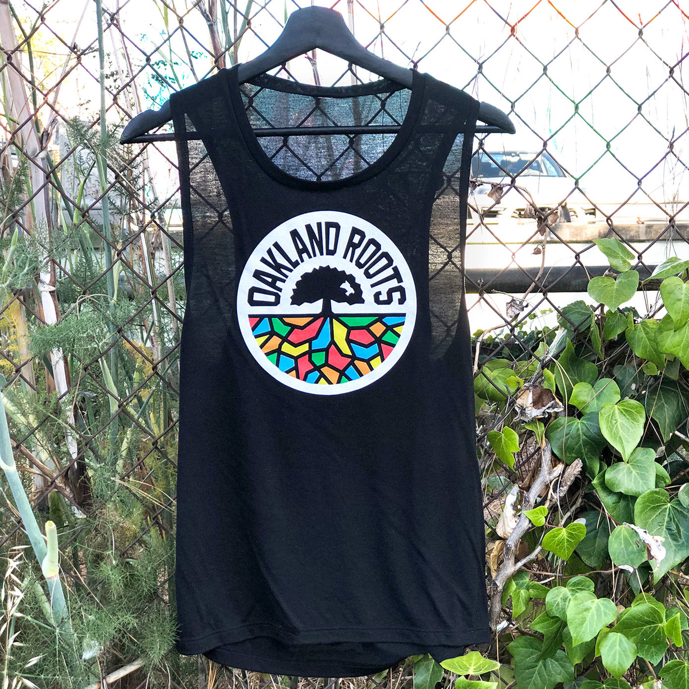 Women’s cut black tank top with full-color Oakland Roots circle logo on the chest hanging outdoors on a fence.