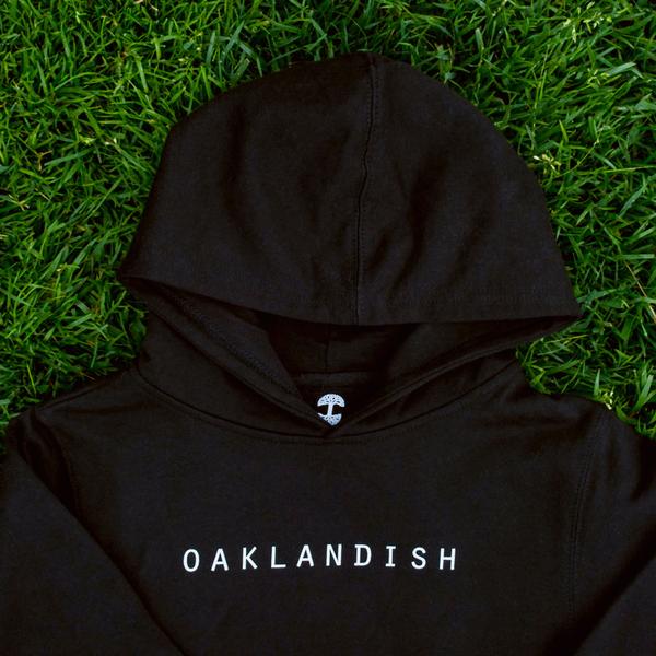 Close-up on top half of a black youth hoodie with white Oaklandish wordmark on the chest, lying on grass.