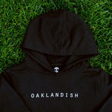Close-up on top half of a black youth hoodie with white Oaklandish wordmark on the chest, lying on grass.