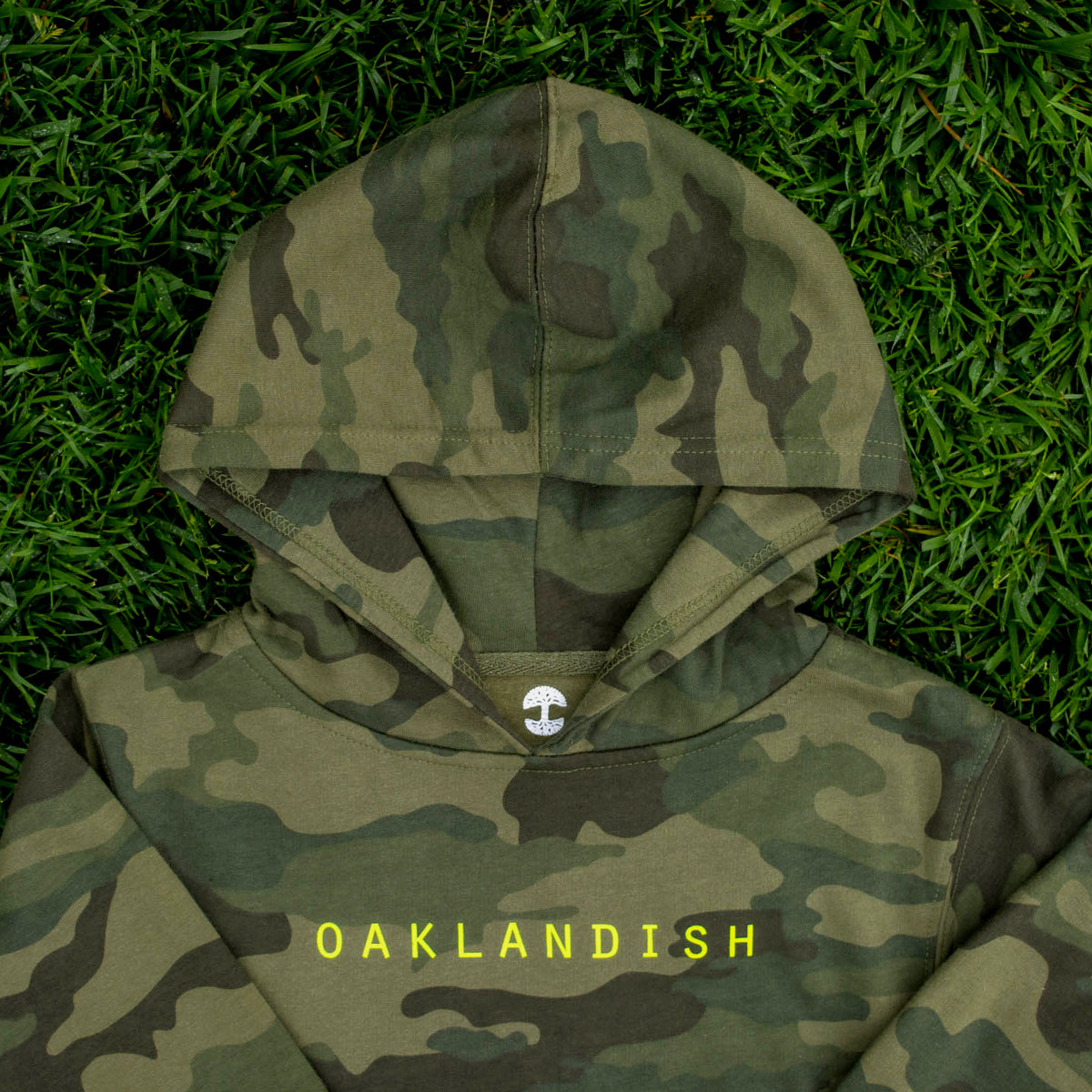 Close up of the top half of a green camo youth hoodie with large green Oaklandish tree logo lying on grass.