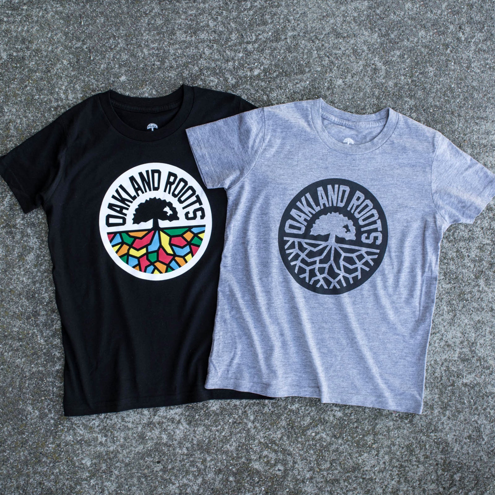 Two youth-sized t-shirts on cement, one black with full-color logo and one grey with black logo.